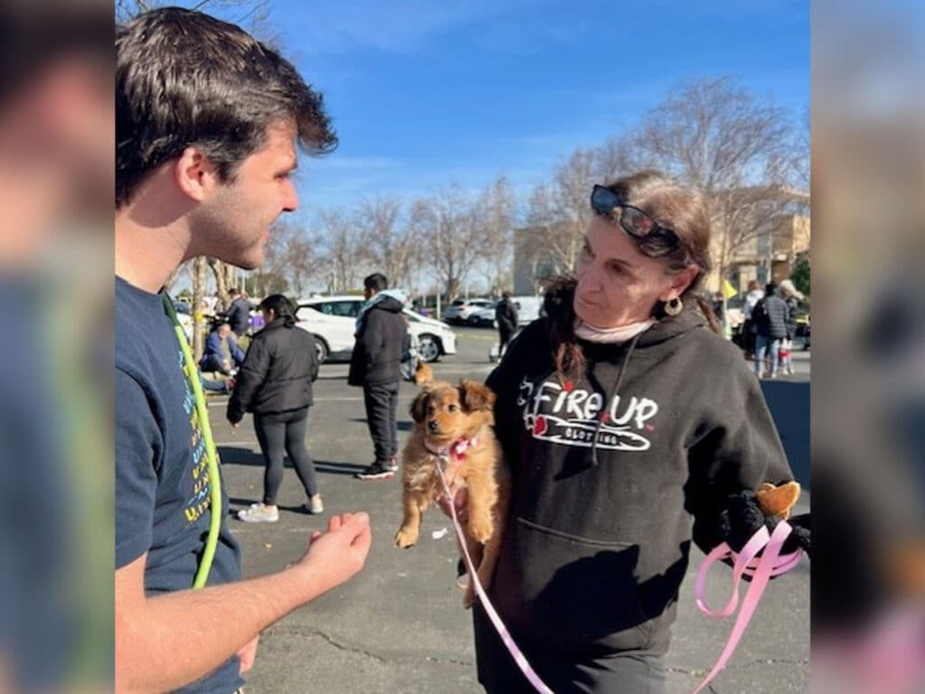 A man in a blue shirt speaks to a woman holding a small brown puppy on a pink leash. The scene is outdoors with cars parked in the background and people waiting in line. Trees are visible against a clear sky.