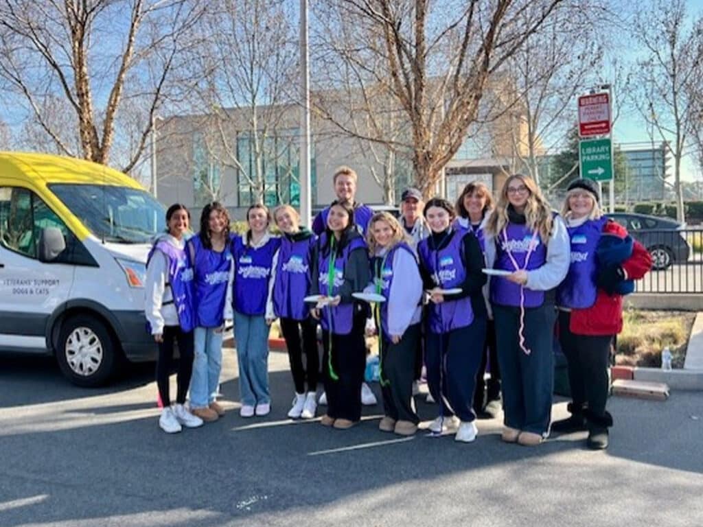 A group of volunteers wearing purple vests stands together on a sunny day outside a building. They smile and hold supplies, with a van parked nearby. The scene portrays a community event related to health services and support.
