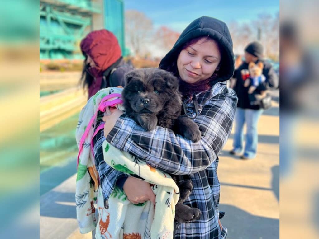 A person wearing a hooded jacket holds a small black dog wrapped in a colorful blanket. In the background, several people are standing in line, dressed in winter clothing, and a building and trees are visible.
