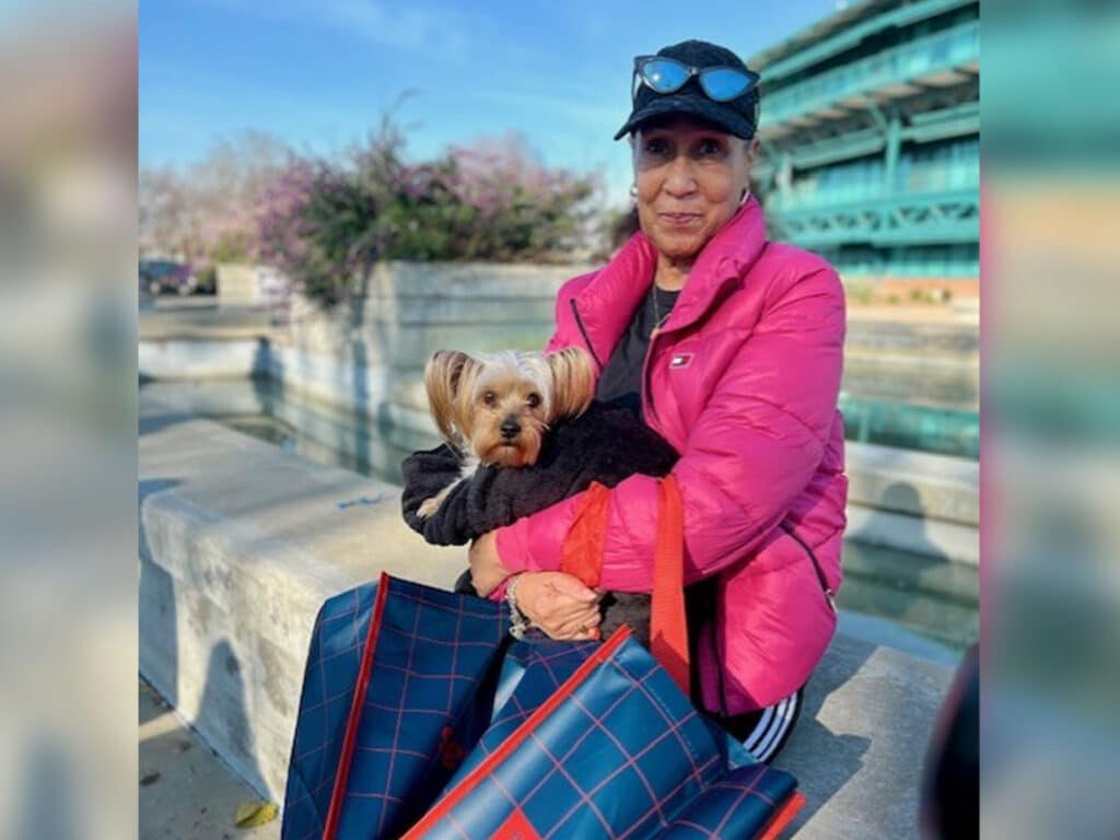 A woman wearing a bright pink jacket and a baseball cap holds a small dog on her lap while sitting on a stone bench. Several blue and red patterned bags are beside her, with greenery and a blurred background featuring flowers and structures.