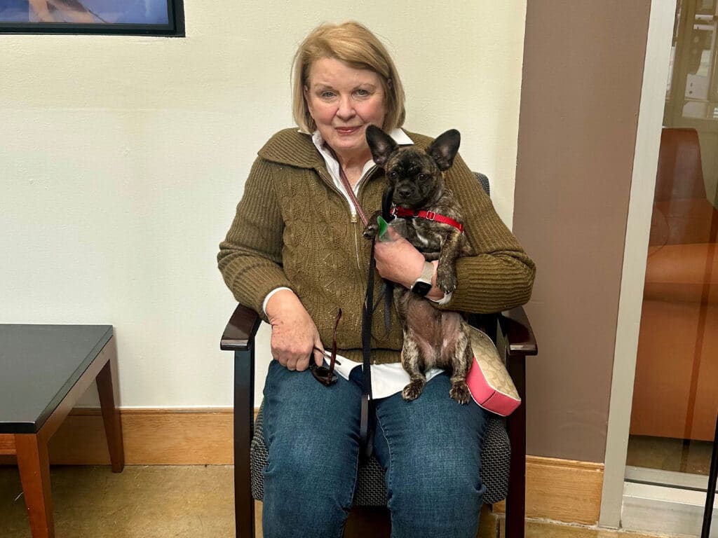 An older woman sitting in a chair, holding a small black dog with a red collar. The woman wears a brown sweater and blue jeans. A side table is visible next to her, and there is a light-colored wall in the background.