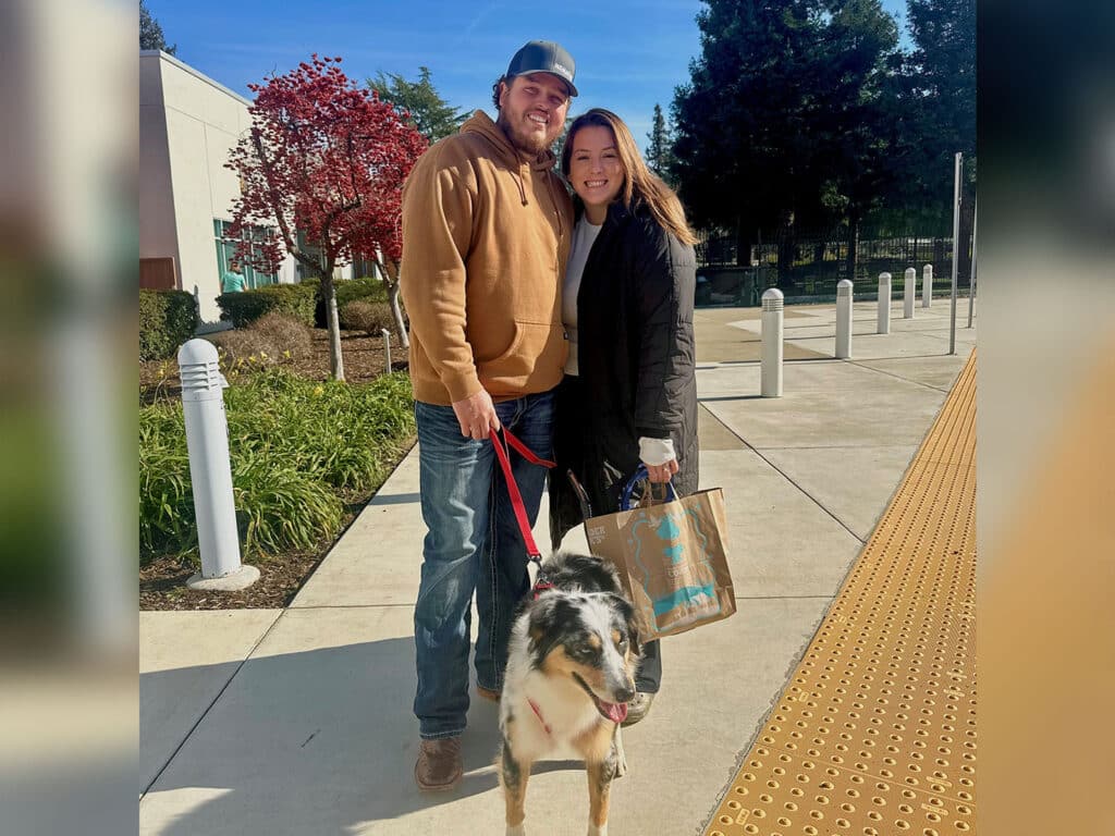 A couple stands outside a building, smiling while holding a leash attached to a dog. The man is wearing a brown hoodie and jeans, while the woman is dressed in a black coat. They have a shopping bag in hand, with green shrubs and trees in the background.