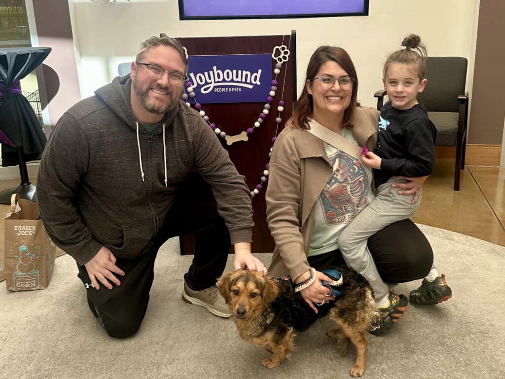 A family poses in a welcoming indoor space. A man and woman kneel beside a small brown dog, while a child sits on the woman's lap. Behind them is a decorative banner that reads "Joybound for Pets".
