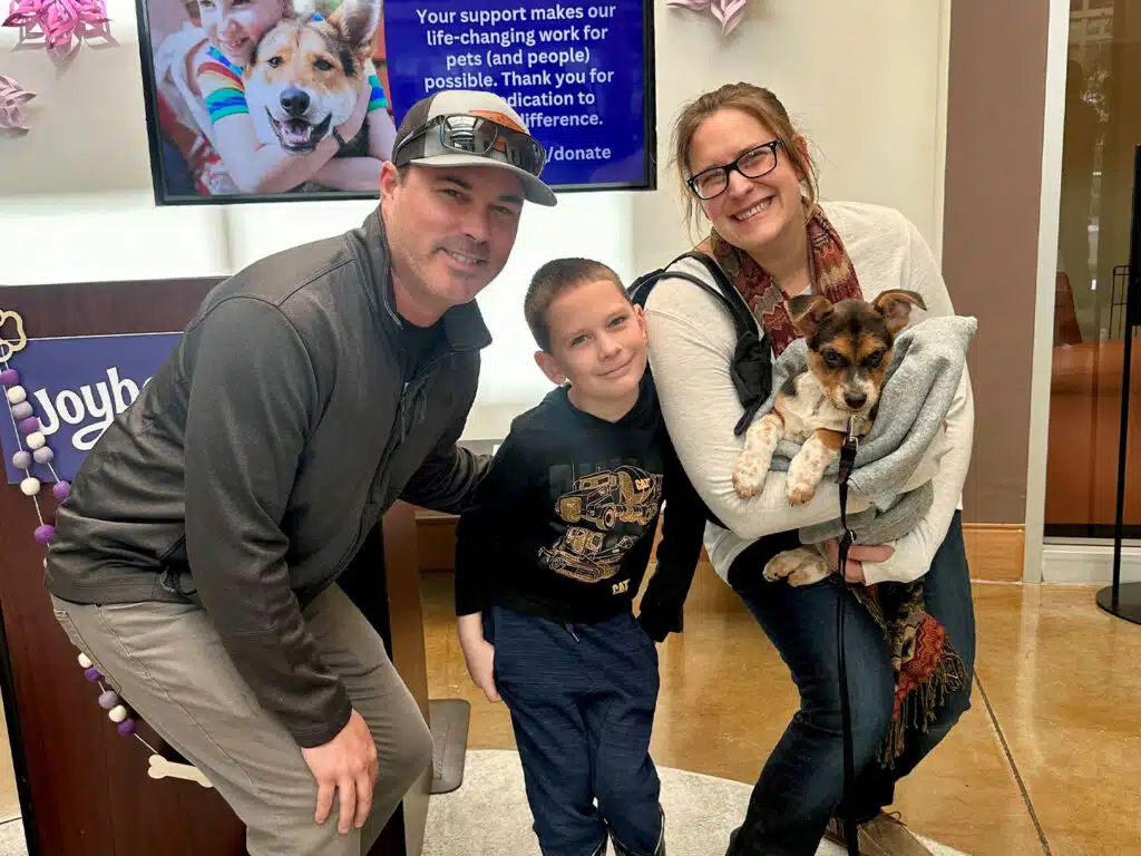A man and a woman, each holding a small dog, pose with a young boy at an adoption event. They are smiling in a brightly lit room with a supportive message displayed on a screen in the background.