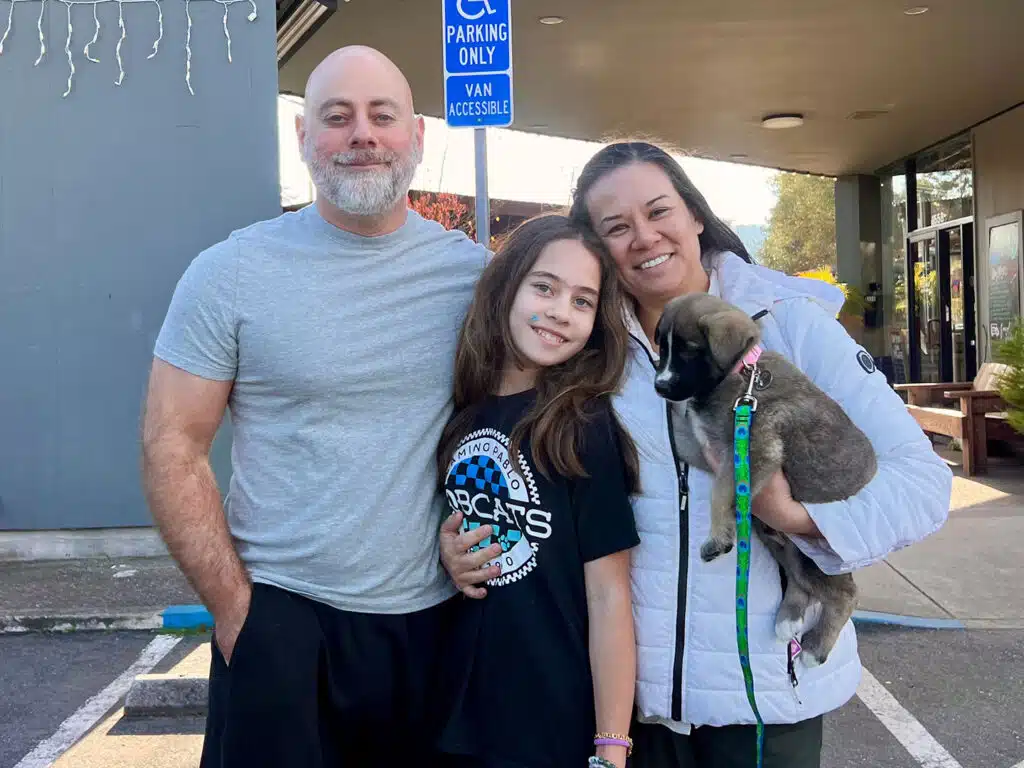 A family of three stands outside a building. A father and mother, both casually dressed, flank their daughter, who is holding a small dog. The parents smile at the camera, and the daughter looks happy. A parking sign is visible in the background.