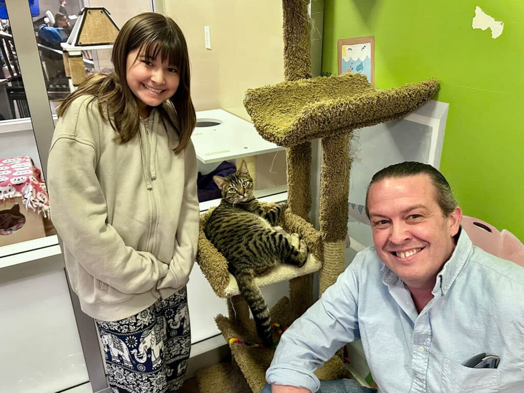 A young girl and a man smiling beside a cat resting on a scratching post inside an adoption center. The girl wears a light gray hoodie and patterned pants, while the man is in a light blue shirt. Brightly colored walls and a playful atmosphere are visible.