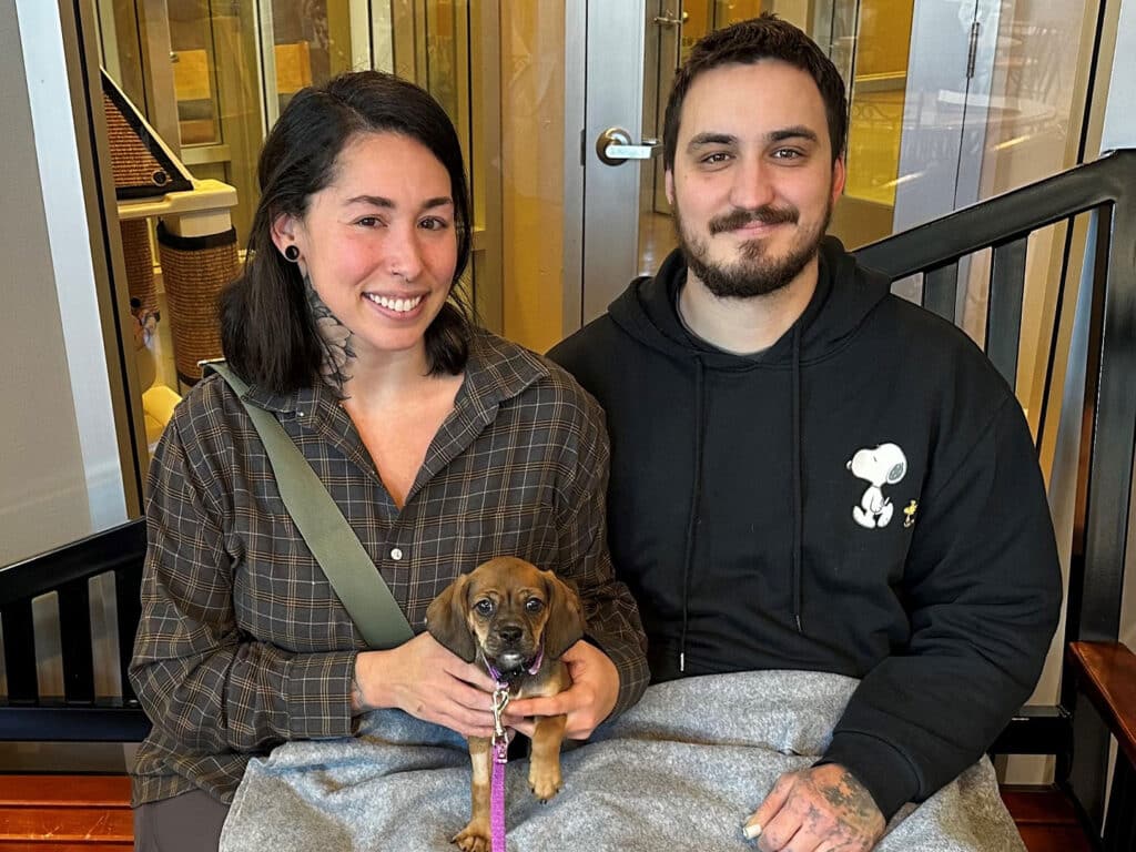 A woman and a man sitting together on a bench hold a small brown puppy. The woman, wearing a plaid shirt, has dark hair and tattoos, while the man, in a black hoodie, has short dark hair. They are indoors, with a modern setting in the background.