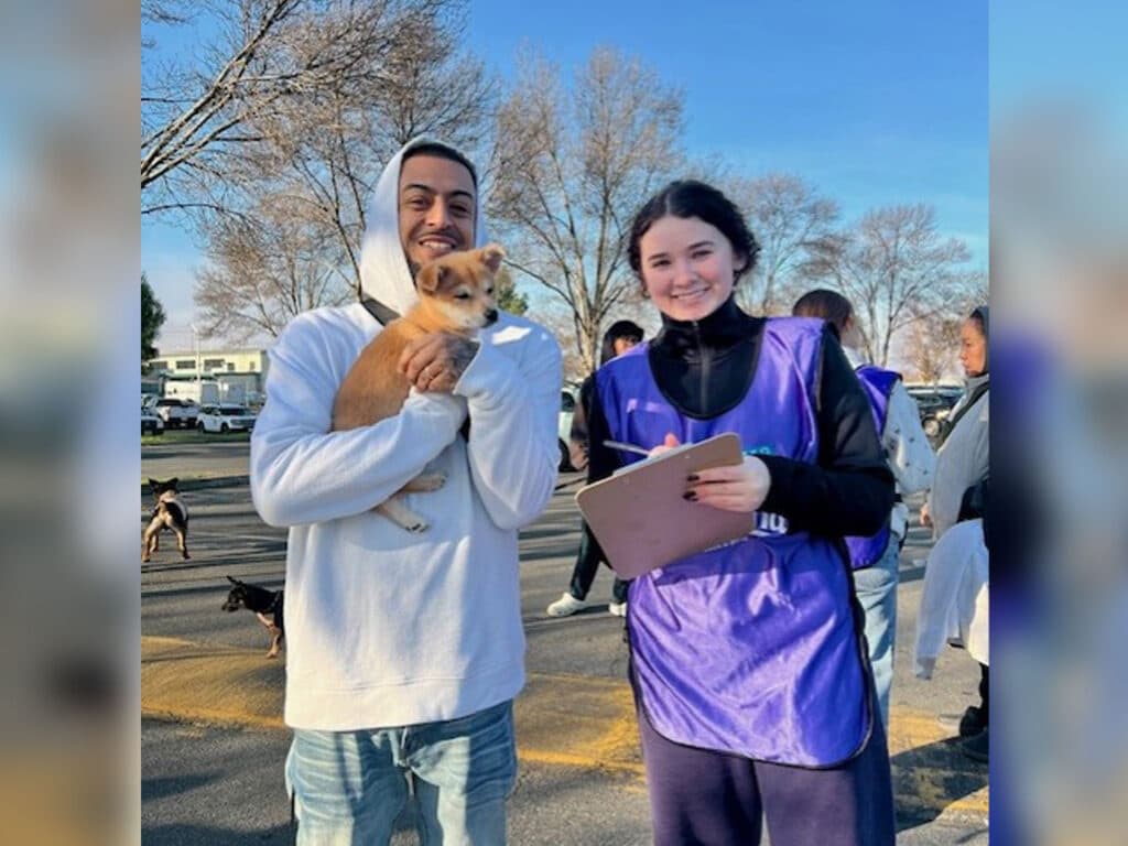 A young man holding a small dog smiles while standing next to a young woman in a purple vest, who is holding a clipboard. They are outdoors with trees in the background, and several dogs are visible on the ground.