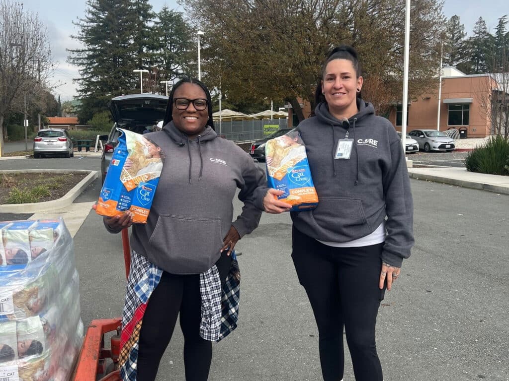 Two women stand in a parking lot, smiling while holding bags of food. Both are wearing gray hoodies, with one woman additionally wearing a checkered shirt around her waist. In the background, parked cars and a building are visible.