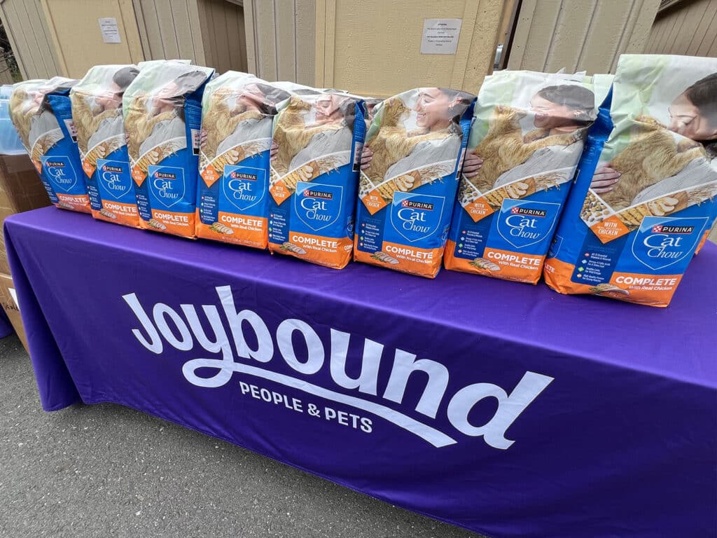 Several bags of Purina Cat Chow, prominently displayed on a purple tablecloth with the words "Joybound People & Pets". The bags are sealed and show images of cats, emphasizing the pet food being offered at the FoodShare Pantry event.