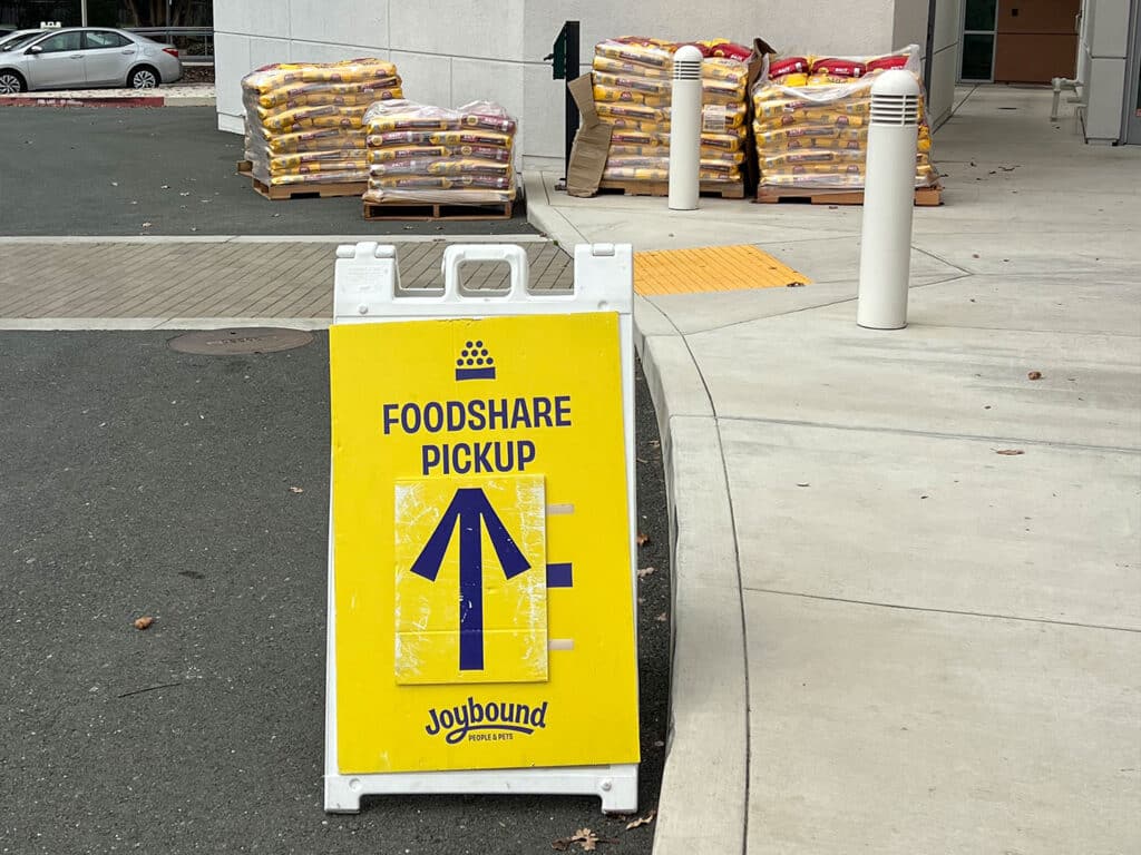A yellow A-frame sign displaying "FOODSHARE PICKUP" with an upward arrow is placed on a paved pathway. In the background, bags of groceries are stacked on pallets outside a building.