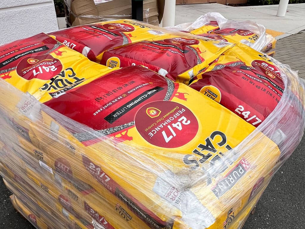 A stack of bags of cat food, prominently labeled with a red and yellow design, stacked on a pallet. The bags display the brand name and weights, indicating they are intended for a pet food pantry. The setting is outdoors, on a paved surface.