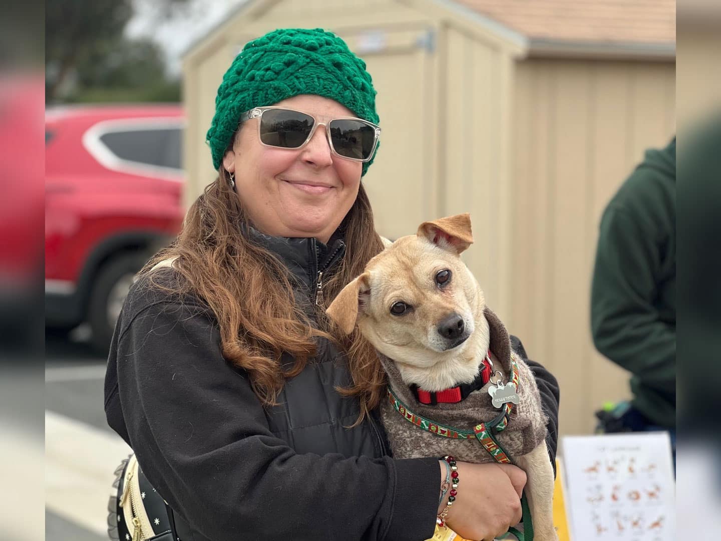 A woman wearing a green knit hat and sunglasses smiles while holding a small dog with a light brown coat. They are outdoors, with a blurred background of vehicles and a building.