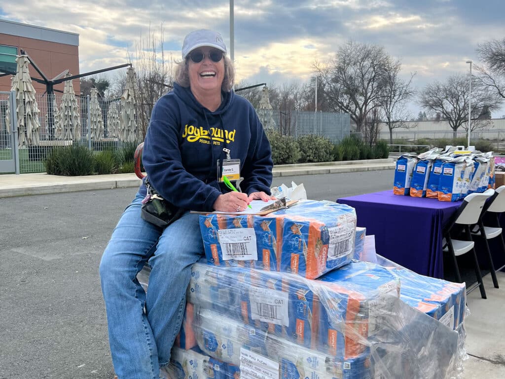 A woman wearing a blue cap and sunglasses sits on a stack of boxes, smiling and writing on a clipboard. The setting has trees and a table with food items in the background, indicating an outdoor food distribution event.