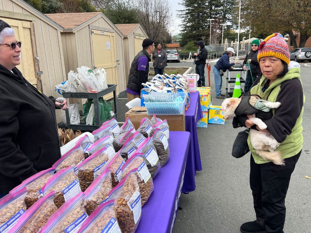 A woman with a colorful knitted hat holds a cat while speaking to another woman at a community food pantry. In the foreground, bags of dry food are arranged on a purple table. Several people are visible in the background near storage sheds.