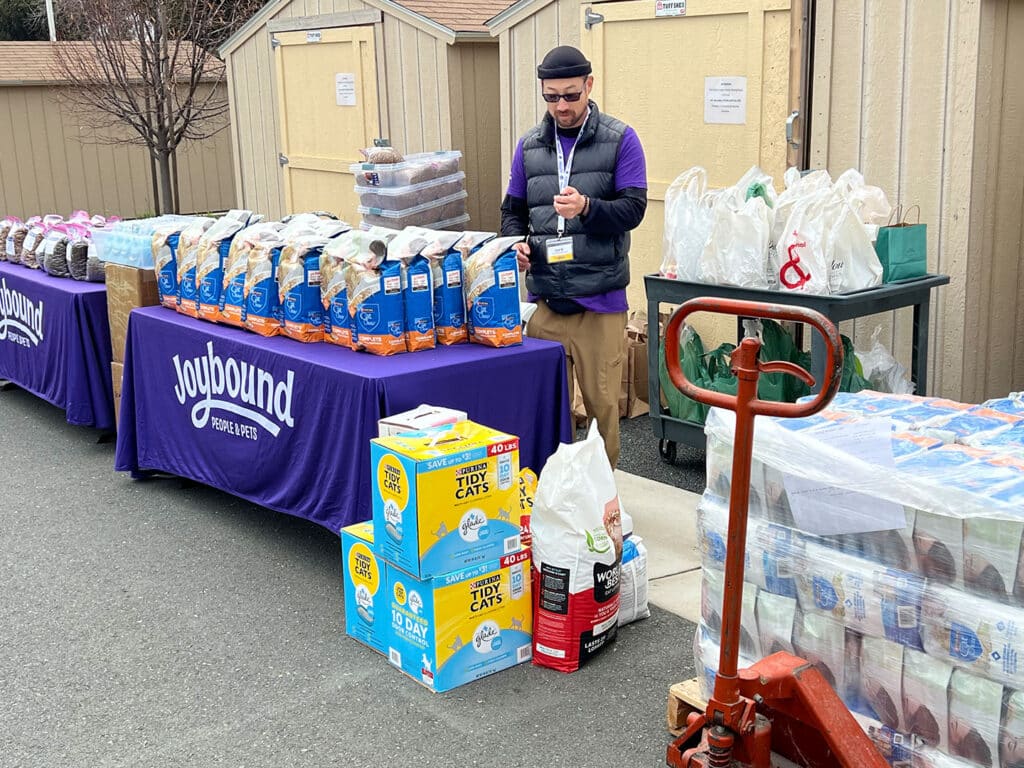 A man in a purple Joybound shirt stands beside a table covered with food items, including bags of snacks and drinks. The background features storage sheds, and a cart is partially visible with additional supplies.