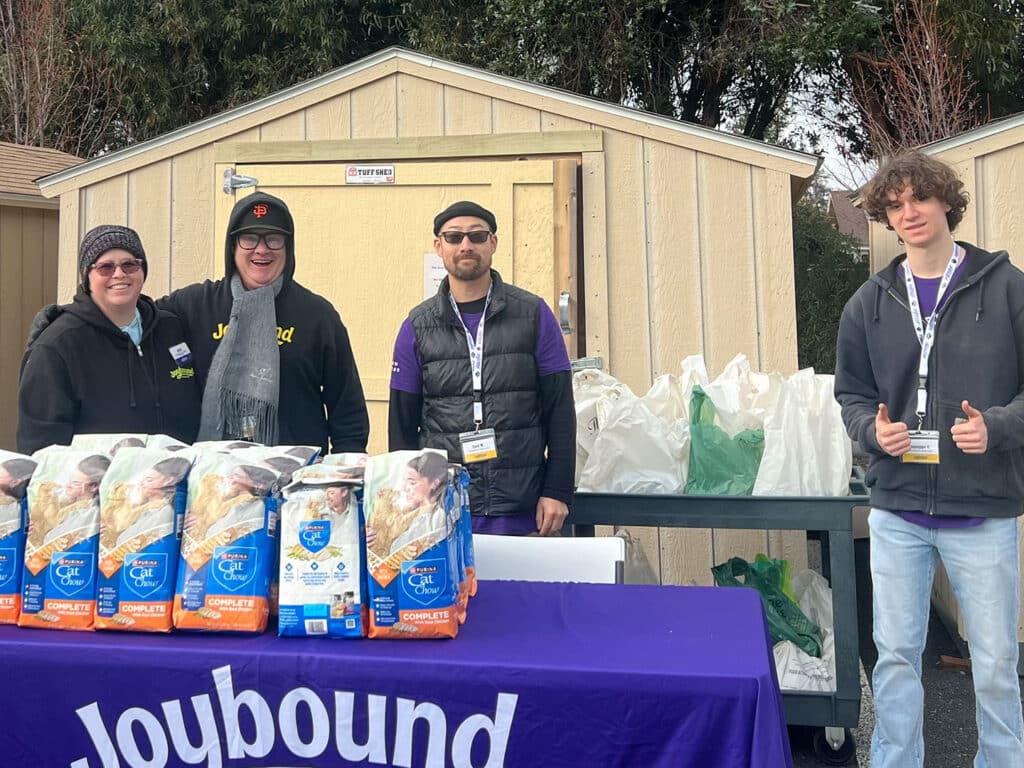 Four individuals stand behind a table covered with bags of food labeled 'Cat Food.' They are in front of an outdoor storage shed, with a few green bags in the background. All are wearing name badges and casual attire.