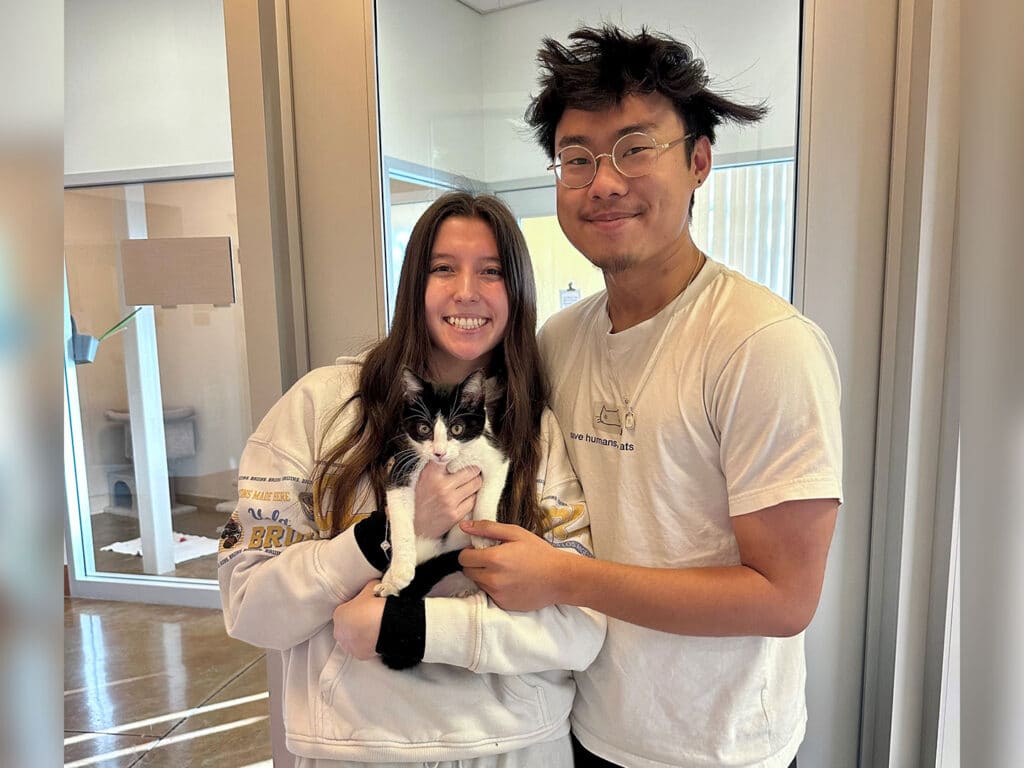 A young woman and a young man stand together in a bright room with large windows, holding a black and white cat. The woman smiles while wearing a sweatshirt, and the man wears a light-colored t-shirt and glasses.