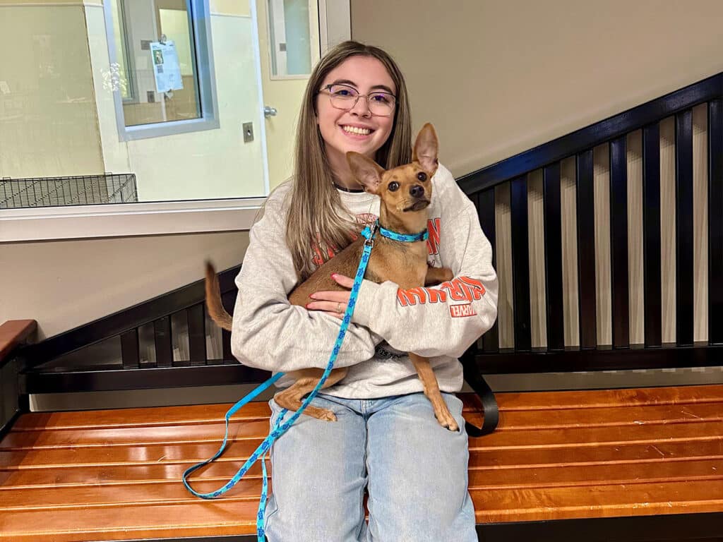 A young woman with long hair and glasses smiles while sitting on a wooden bench, holding a small brown dog with large ears. The dog is secured with a leash, and they are in a brightly lit indoor space.