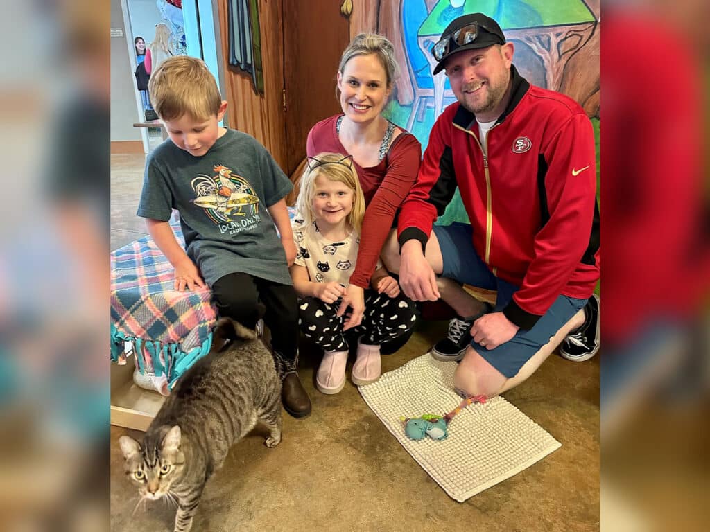 A family poses inside an animal shelter. A woman and a man kneel beside two children, one boy and one girl, who both smile. A gray tabby cat sits on a mat in front of them. The background features colorful mural artwork and animal care items.
