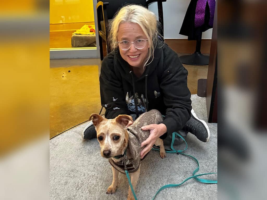 A woman with glasses and long blonde hair kneels on a gray carpet, gently petting a small brown dog with a harness. The background shows a softly lit room with pet supplies, conveying a calm atmosphere.