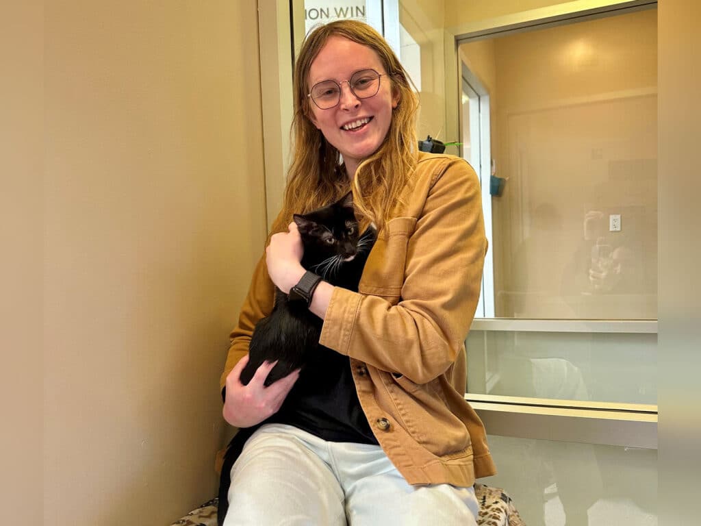 A person with long hair and glasses sits in a cozy room, holding a black cat in their arms. The individual is wearing a tan jacket and a black shirt, smiling while surrounded by light-colored walls and windows.