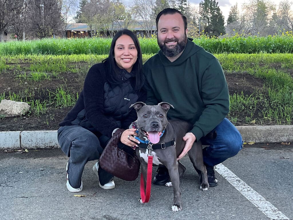 A woman and a man are smiling while sitting on the ground beside a gray dog with a pink tongue, in a grassy area with trees in the background. The woman is wearing a black jacket and the man is in a green hoodie.