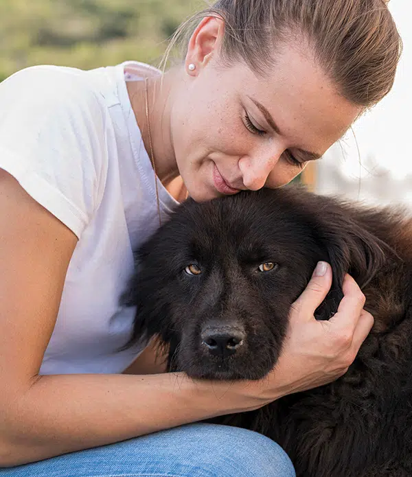 A woman sits close to a black dog, gently hugging its head while resting her face against it. They are outdoors, surrounded by greenery. This image represents the bond between humans and emotional support animals.