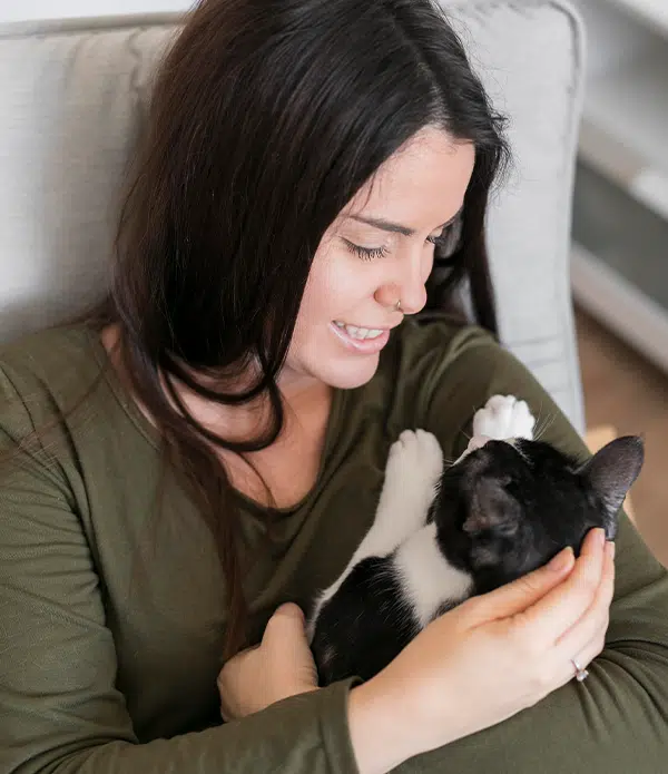 A woman sitting on a sofa, smiling gently at a black and white cat cradled in her arms. The cat appears relaxed, with its paws extended, while the woman has long dark hair and is wearing a green top. This image highlights the bond between humans and emotional support animals.