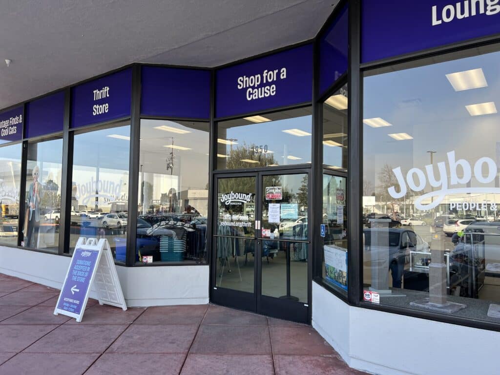 Exterior view of Joybound thrift store with large glass windows displaying the store name and promotions. A sidewalk sign reads 'Shop for a Cause.' Parked cars are visible in the background, emphasizing a community shopping environment.