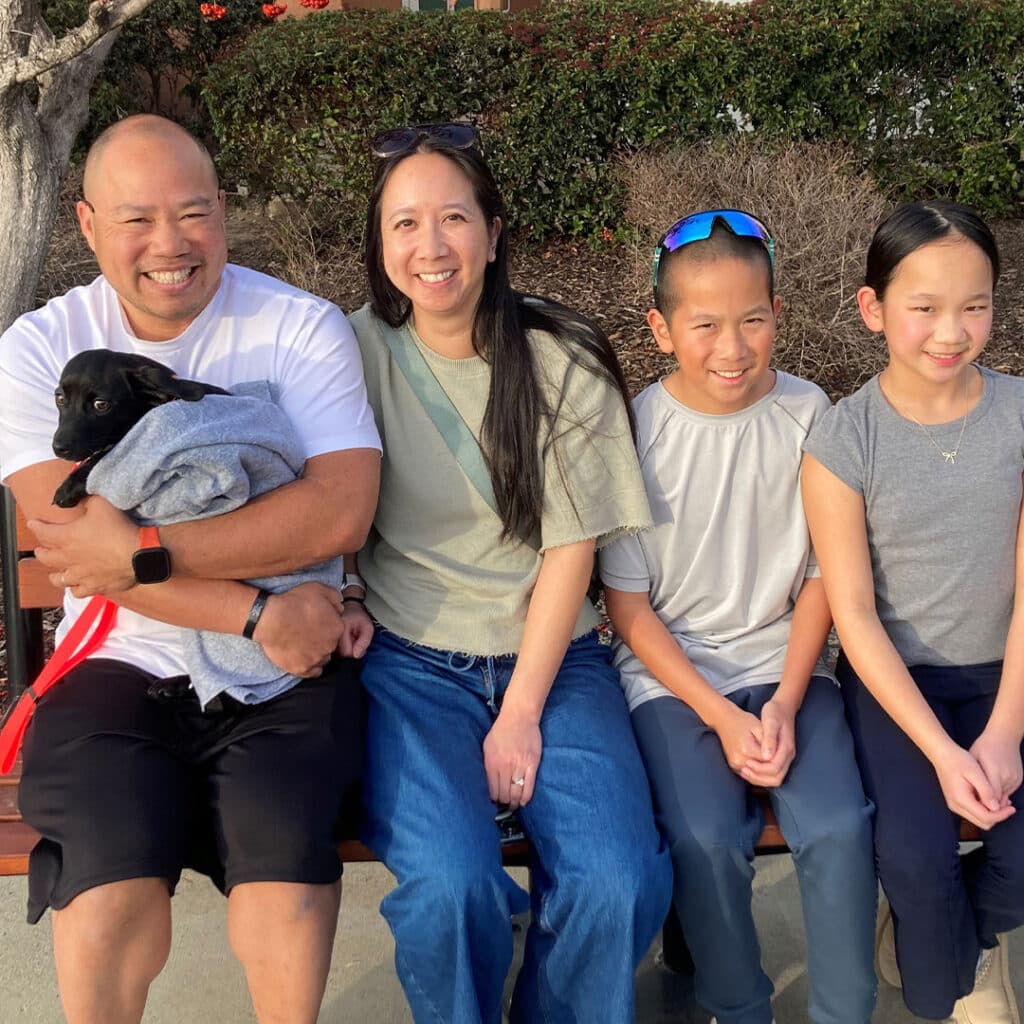 A family sitting on a bench outside. A man holds a small black dog wrapped in a blanket, while a woman sits beside him. Two children, a boy and a girl, sit next to them, all smiling. The background includes greenery and a pathway.