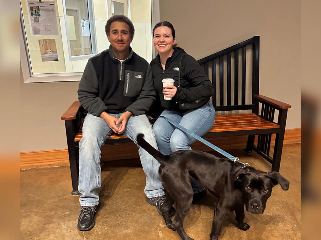 A man and a woman sitting on a wooden bench in a room. The woman is holding a coffee cup, and a black dog with a leash is standing nearby. A window is visible behind them, providing light to the space.