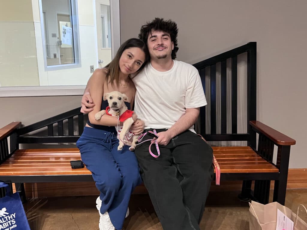 A man and a woman sitting on a wooden bench inside an adoption center, smiling. The woman holds a small dog in a red sweater, while a bag is placed on the floor nearby. The setting shows a cozy, welcoming environment.