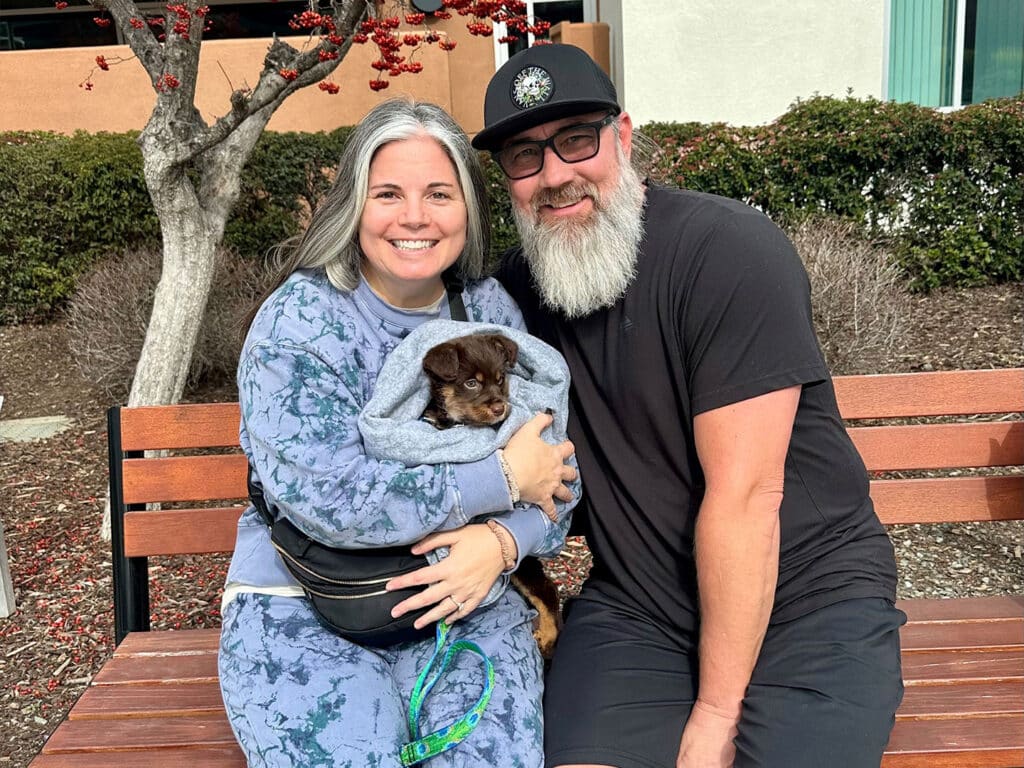 A woman and a man sit on a bench outdoors, smiling while holding a small dog wrapped in a blanket. The woman has gray hair and is wearing a blue outfit, while the man has a beard and wears a black t-shirt and a cap. They are surrounded by greenery.