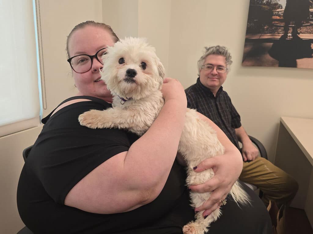 A woman with glasses holding a small white dog on her lap, smiling at the camera. In the background, a man with gray hair is seated, looking towards the woman and dog. The setting appears to be an office with neutral-colored walls.