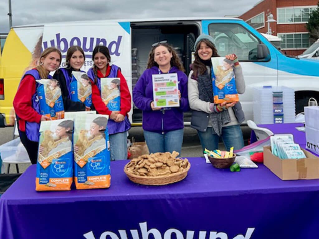 A group of five volunteers standing behind a table filled with food bags and cookies at a community event. The table has a purple tablecloth with the word "Inbound" and displays additional items. A blue and yellow van is parked in the background.
