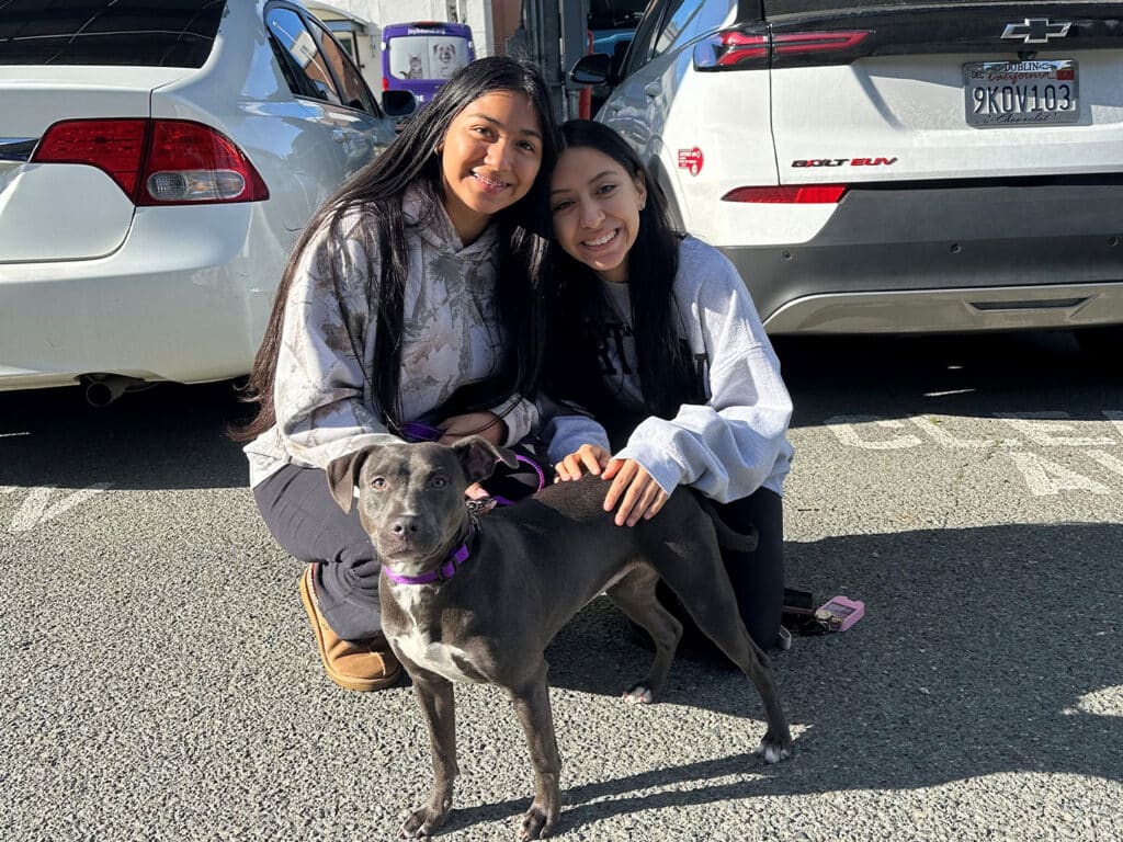 Two young women crouched on a parking lot beside a grey dog wearing a purple collar. They are smiling at the camera, with one woman wearing a grey sweatshirt and black pants, and the other in a white sweatshirt. A parked car is visible in the background.
