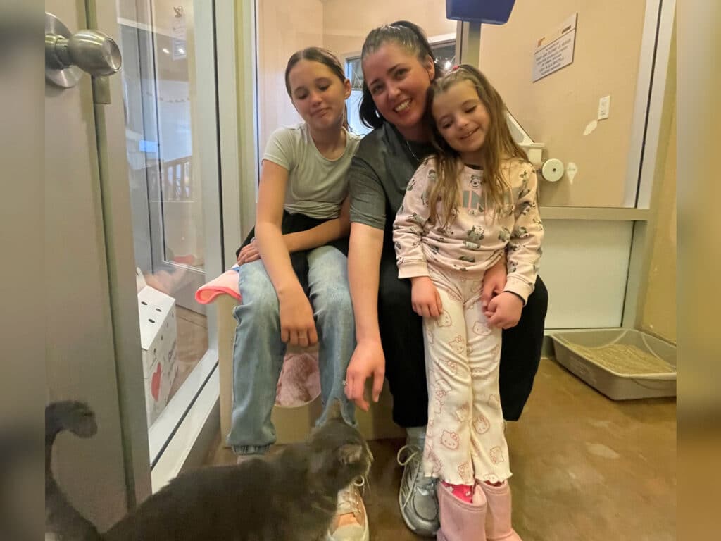 A woman sits on a bench inside a pet adoption facility, smiling as she interacts with two young girls next to her. One child pets a gray cat sitting in front of them. The setting is bright and inviting, with cat bedding and adoption information visible.