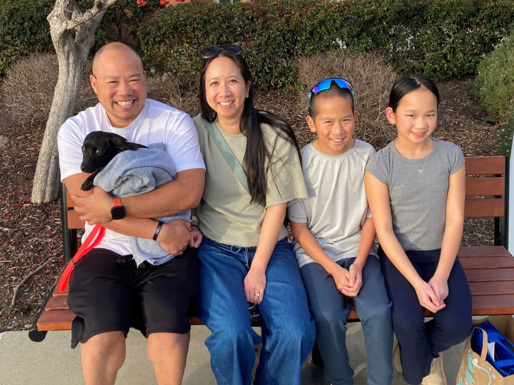 A family of four sitting on a bench outdoors. The man on the left is holding a small black puppy wrapped in a blanket. The woman next to him smiles, along with two children, a boy and a girl. They are all casually dressed, surrounded by greenery.