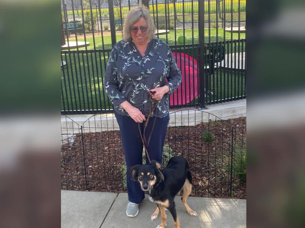 A woman stands outside a fenced area holding a leash attached to a dog. She wears a floral blouse and dark pants, smiling while looking at the camera. The background shows a green yard with a pink object visible.
