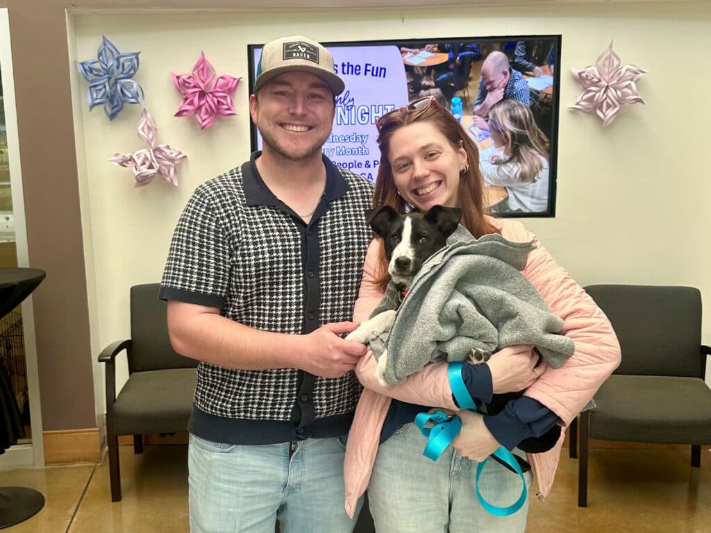 A man and woman stand together smiling, holding a small dog wrapped in a gray blanket. The woman has long brown hair and wears a pink jacket, while the man wears a black and white patterned shirt and a cap. In the background, decorations and a screen are visible.