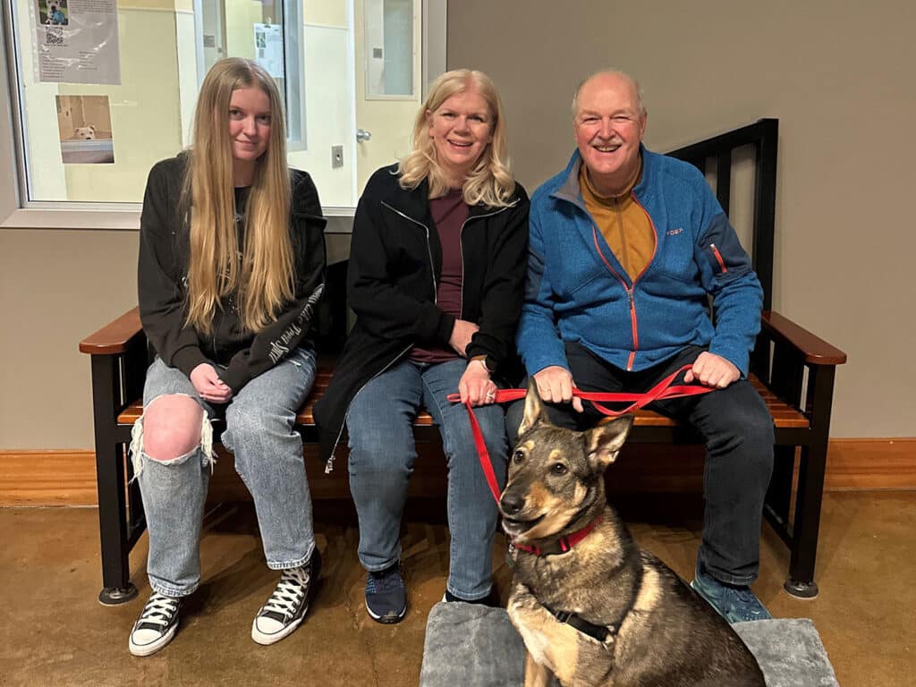 A teenage girl, a woman, and a man sit on a wooden bench with a dog on a leash in front of them. The dog, a German Shepherd mix, sits on a mat. The setting appears to be an indoor area, possibly at an animal shelter.