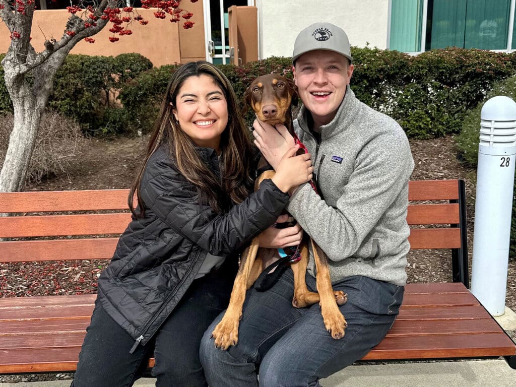 A woman and a man sitting on a wooden bench, each holding a brown dog with floppy ears. The woman is smiling and wearing a black jacket, while the man is in a gray sweater and cap. Behind them, there are some shrubs and part of a building.