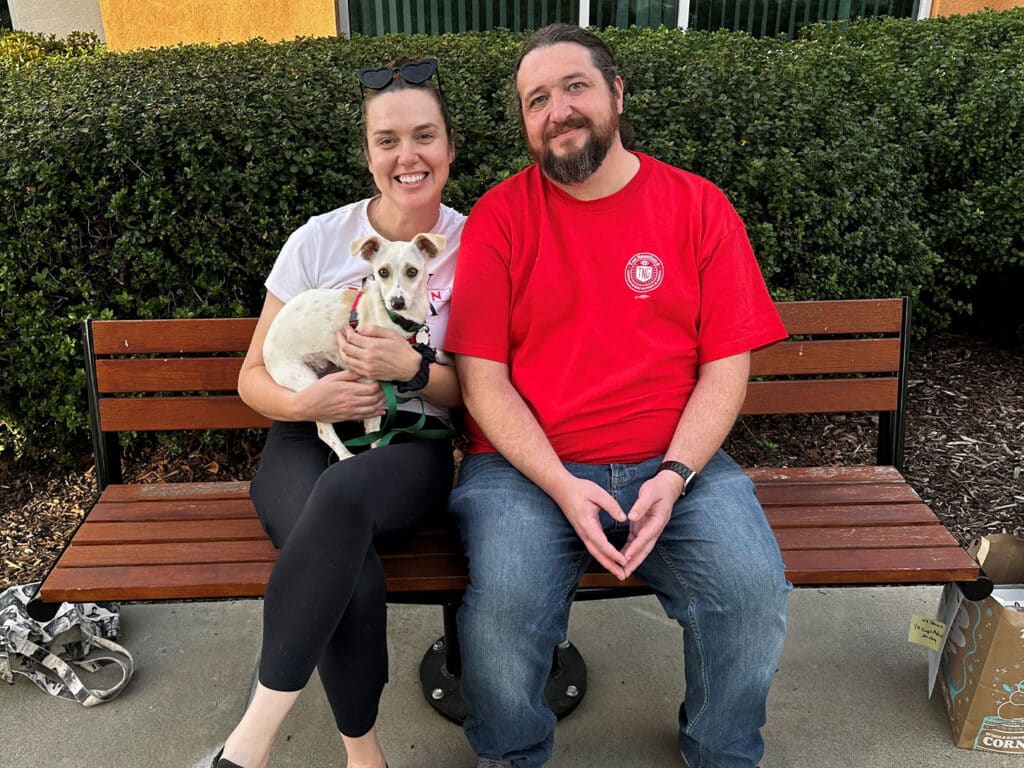A woman with dark hair wearing a white t-shirt and a man with light brown hair in a red t-shirt sit together on a bench, holding a small white dog. They are outdoors with green shrubbery in the background.