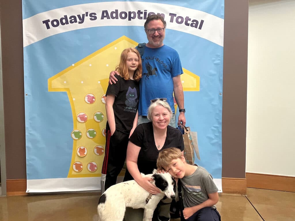 A family of four stands in front of a colorful backdrop that reads 'Today's Adoption Total.' The group includes two children, a woman, and a man, with a white dog at their feet. The setting appears to be an animal adoption event.