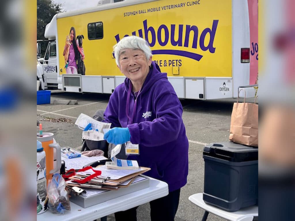 A woman in a purple jacket smiles while standing at a table filled with supplies, next to a mobile veterinary clinic that reads 'Joybound.' The setting appears to be an outdoor event, with parked vehicles and a sunny sky in the background.