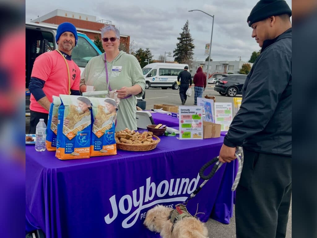 A man in a pink shirt and a woman wearing glasses smile at a booth with a purple tablecloth labeled "Joybound." They display dog food bags and treats, while a man with a dog on a leash approaches. Background includes vehicles and trees.