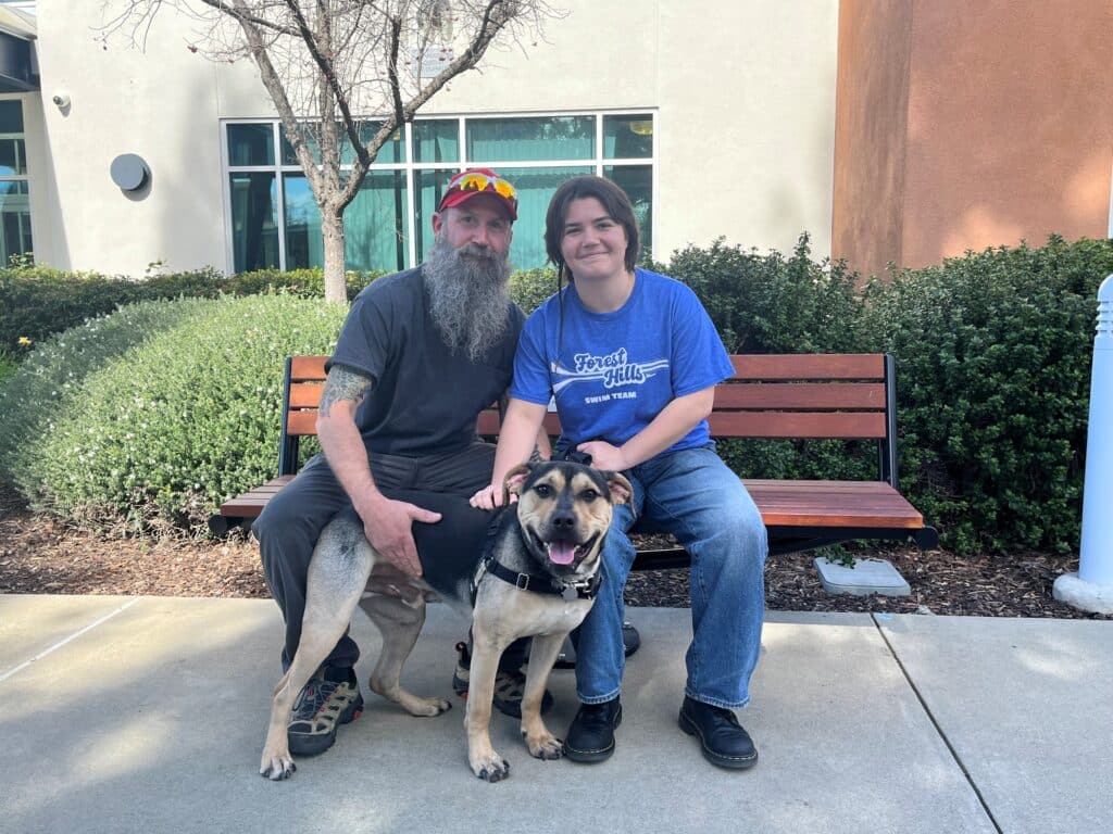 A man with a long beard and a baseball cap sits on a bench alongside a teenage boy. They are both smiling as they pose with a large dog, which is sitting at their feet. The background shows green shrubs and a modern building, representing community pet adoptions.