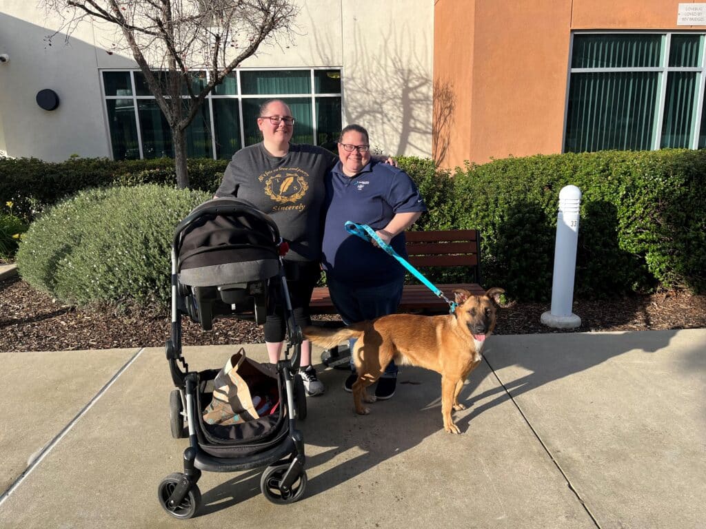 Two women stand outside a building, smiling while holding a dog on a leash and a stroller. One woman wears a gray t-shirt, and the other is in a blue shirt. A green hedge is visible in the background. This image relates to pet adoptions.