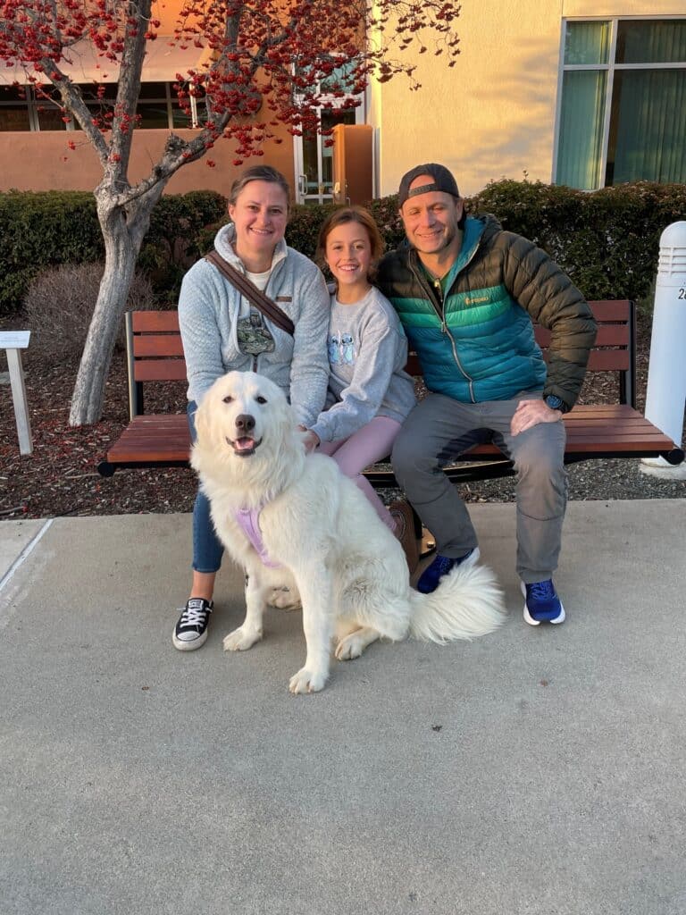 A family of three sits on a bench outside, posing with a fluffy white dog wearing a purple harness. The adults are smiling, while the young girl is excited. Surrounding them are flowering trees, enhancing the cheerful atmosphere. This scene highlights pet adoptions.
